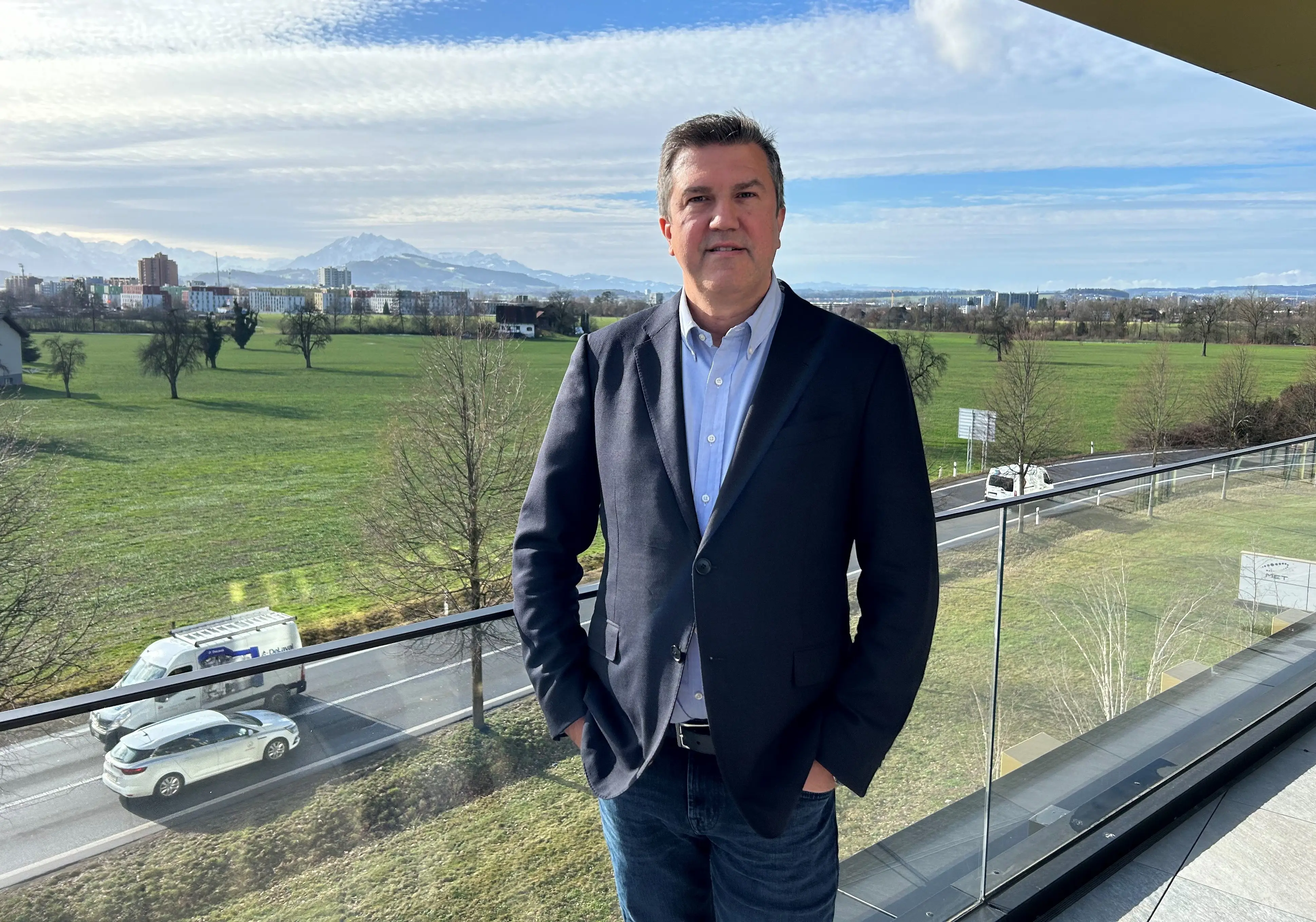 Klaus Reinisch is wearing a dark blazer, light blue button-down shirt, and jeans stands on a balcony overlooking a wide green landscape. In the background, open fields, trees, a roadway with passing vehicles, and distant snow-capped mountains stretch across the horizon under a partly cloudy sky. The glass balcony railing and bright natural daylight create a clear, professional outdoor portrait setting.